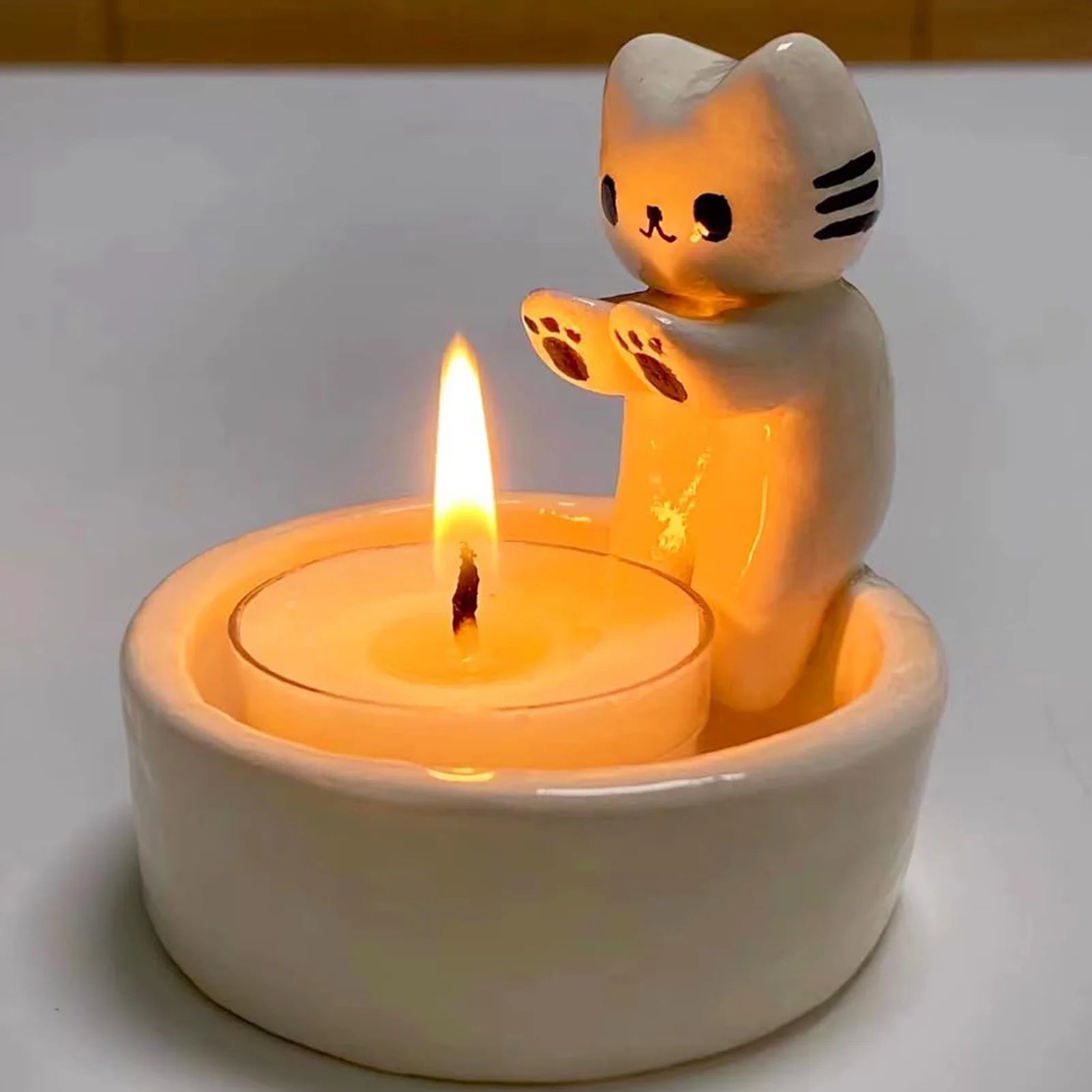 Portavela con gato blanco sentado en el borde de un pequeño cuenco calentando las patas con una vela tipo tea light, sobre fondo blanco